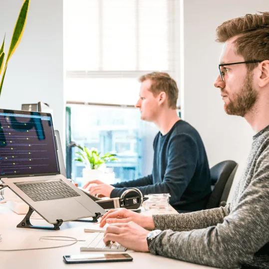 Two men working at computers in a bright office, focused on their tasks. One is in the foreground typing on a keyboard, while the other is in the background facing a monitor—both dedicated to delivering top-notch SEO Kelowna solutions.