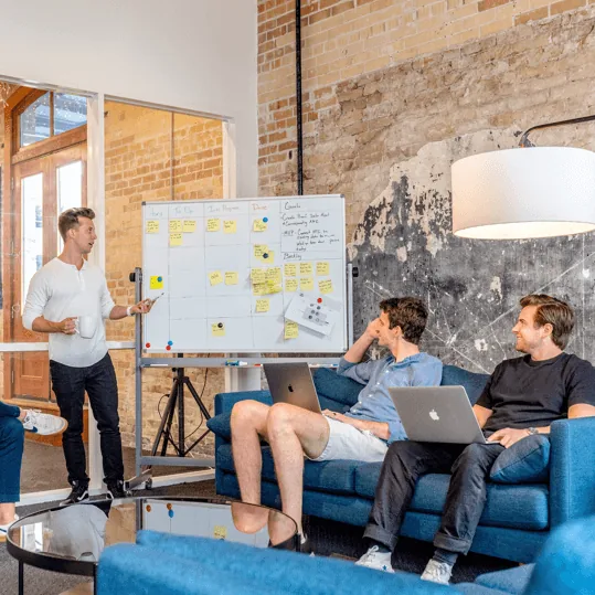 Three people sit on a blue couch with laptops, listening to a man lead an SEO Kelowna presentation by a whiteboard covered with sticky notes in a modern, industrial-style office space.
