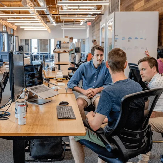 Three men discuss seo kelowna strategies around a desk with computers and laptops in a modern open office. Other people work in the background, while a drink can and keyboard sit on the desk.